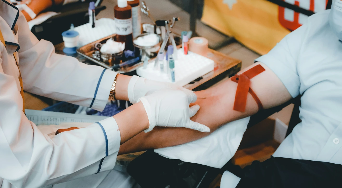 A healthcare worker wearing gloves prepares to draw blood from a person’s arm with a tourniquet on, with sample tubes and supplies on a tray nearby.