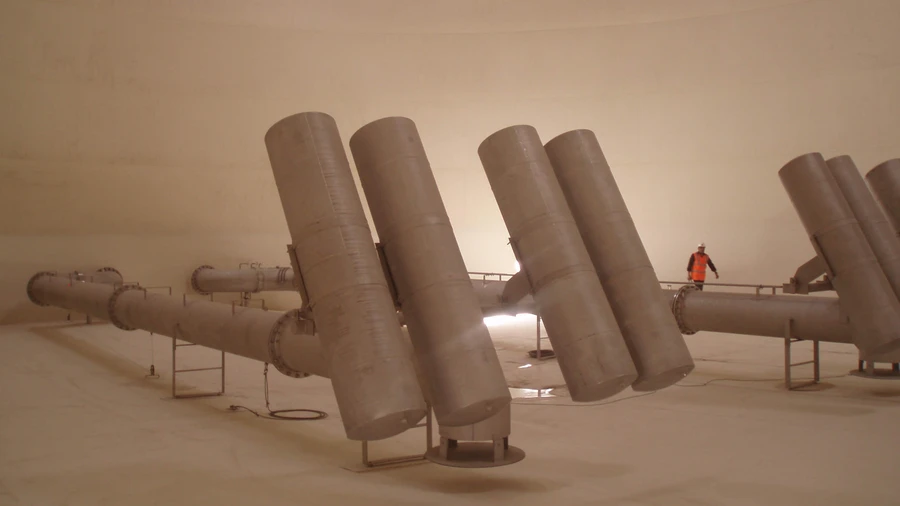 In einem großen Lagertank ragen mehrere große Metallentlüftungsrohre schräg nach oben aus einem Verteiler, während in einiger Entfernung ein Arbeiter in einer orangefarbenen Sicherheitsweste steht.