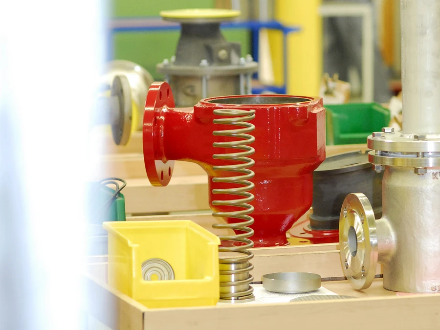 A red industrial valve body and a metal spring sit on a workbench in a factory workshop, with other parts and blurred workers in the background.