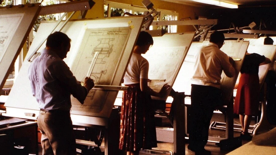 Several people stand at angled drafting tables in a vintage design studio, working on large technical drawings under warm indoor lighting.