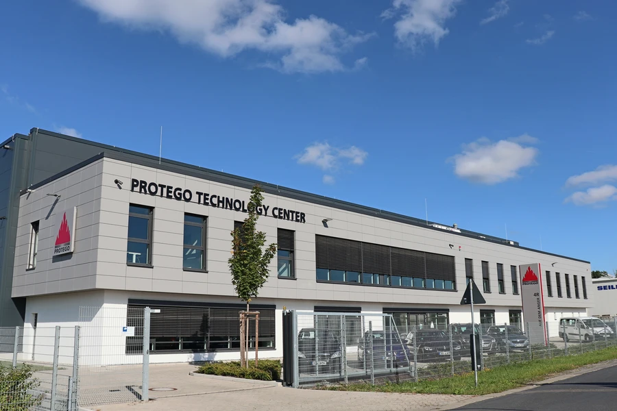 A modern office building labeled “PROTEGO TECHNOLOGY CENTER” under a blue sky, with a fenced parking area in front.