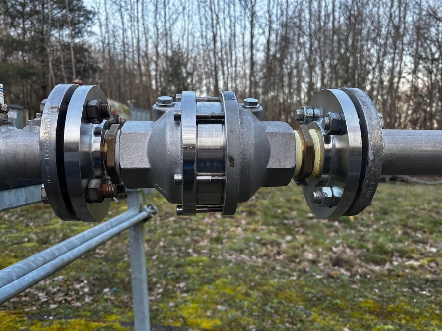 Close-up of a metal pipe coupling with flanges and bolts, installed outdoors with trees in the background.