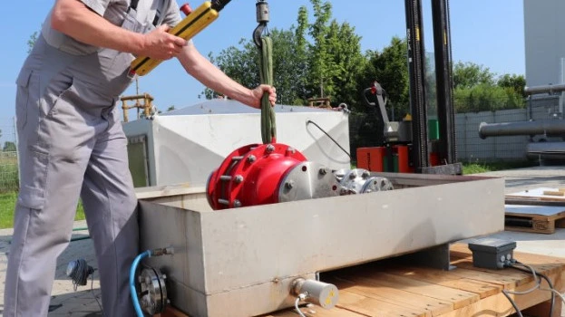 A technician lowers a large red industrial safety device into a rectangular outdoor test basin using a hoist, with cables and piping connected.