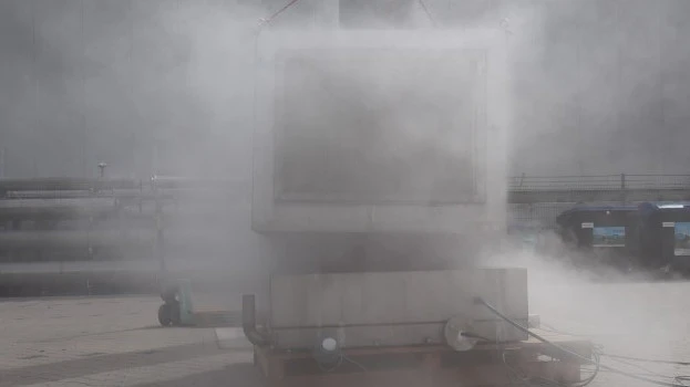 Outdoor industrial test setup with a large container largely obscured by a dense cloud of venting vapor or smoke during an active test.