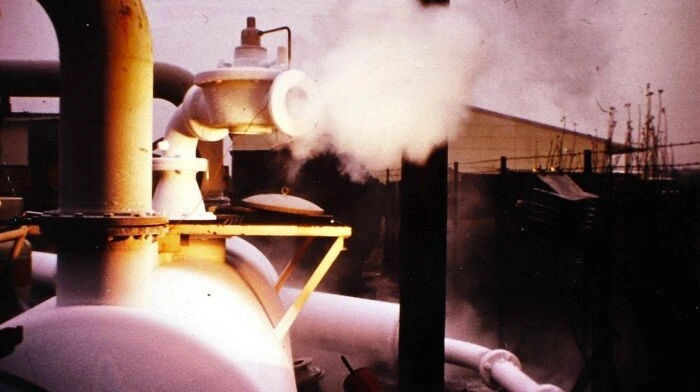 An industrial valve on large outdoor piping releases a plume of white vapor near a facility at dusk.