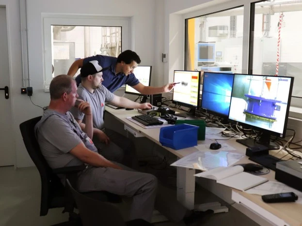 Drei Ingenieure arbeiten gemeinsam an einem Kontrollpult und überprüfen Testdaten und Bilder auf mehreren Computermonitoren in einem technischen Labor.