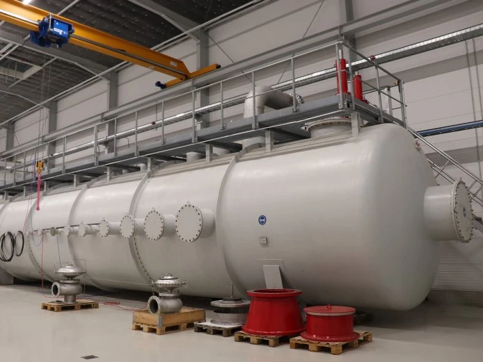 Großer horizontaler Industrie-Druckbehälter in einer Fabrikhalle, ausgestattet mit mehreren Düsen, Ventilen und einer erhöhten Zugangsplattform unter einem Brückenkran.