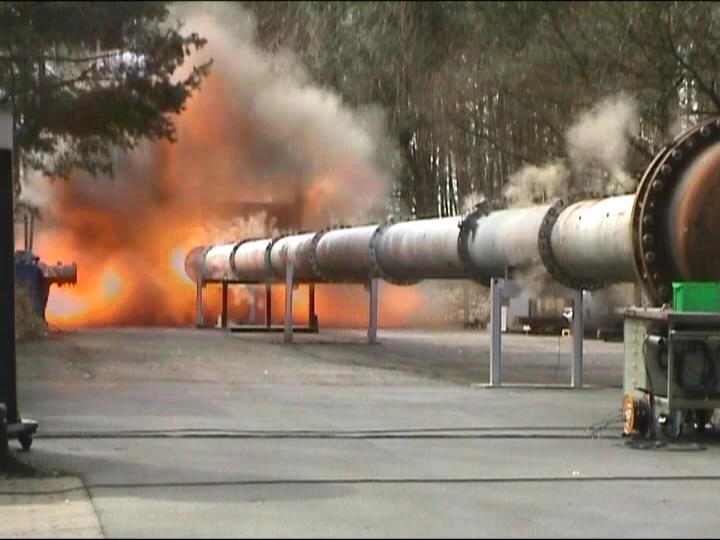 A fireball and thick smoke erupt beside a long elevated pipeline at an outdoor industrial test site.