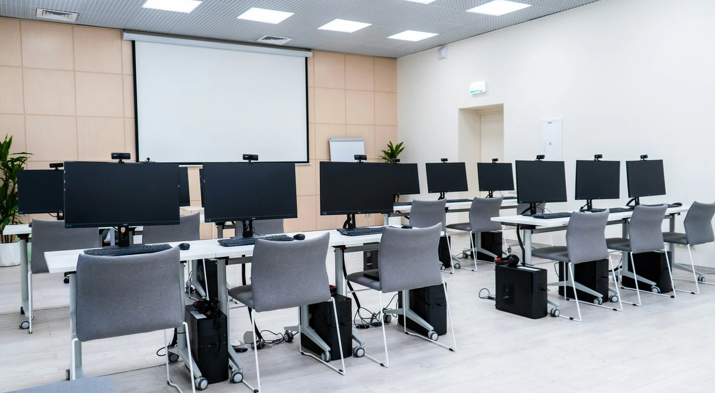 Un aula de informática vacía con filas de pupitres, monitores de escritorio y una gran pantalla de proyección en la parte delantera.
