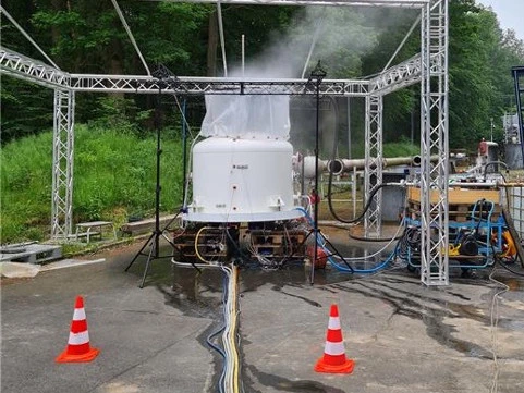 Outdoor industrial test setup with a white vessel venting steam beneath a metal truss frame, surrounded by hoses, equipment, and safety cones.