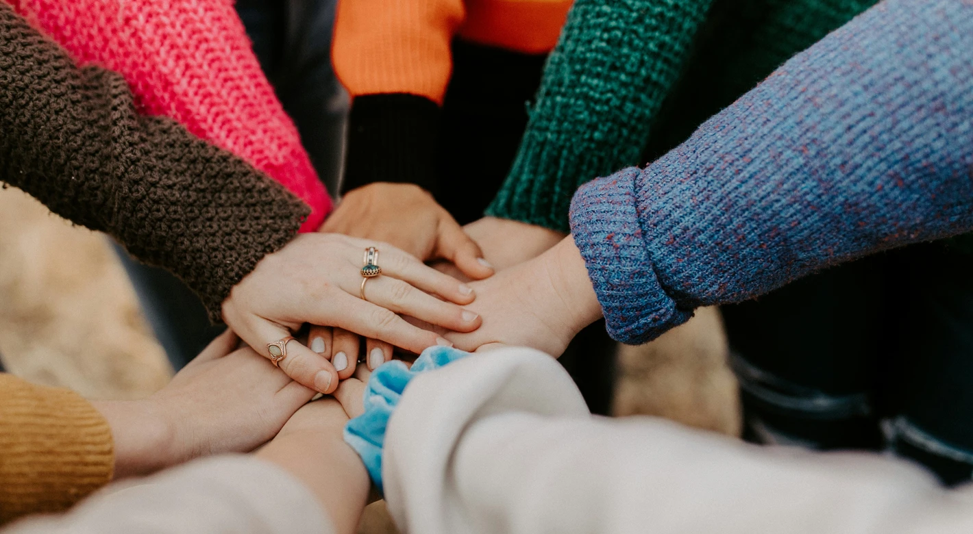 Several people stack their hands together in a team huddle, with colorful sweaters visible around the circle.