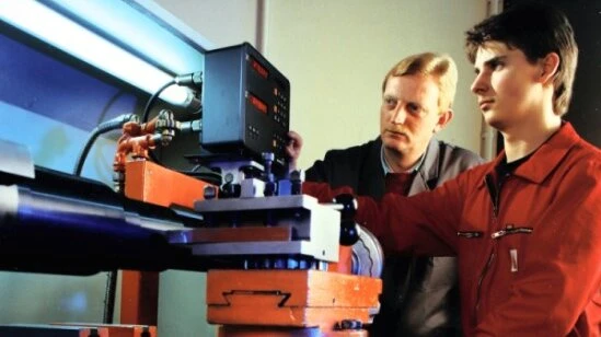 Two technicians operate a large industrial testing machine, with one in red coveralls adjusting the controls while another observes.