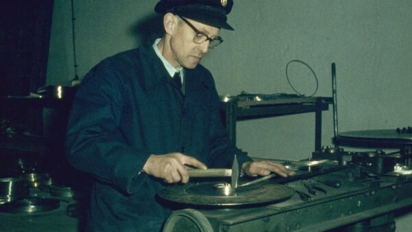 A man in a work coat and cap uses a hand tool at a workbench with metal parts and machinery in a workshop.