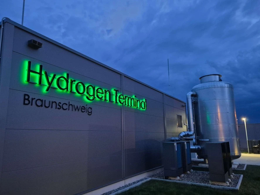 Exterior view of the Hydrogen Terminal Braunschweig building at dusk, with a green illuminated sign and adjacent cylindrical industrial equipment under a cloudy blue sky.