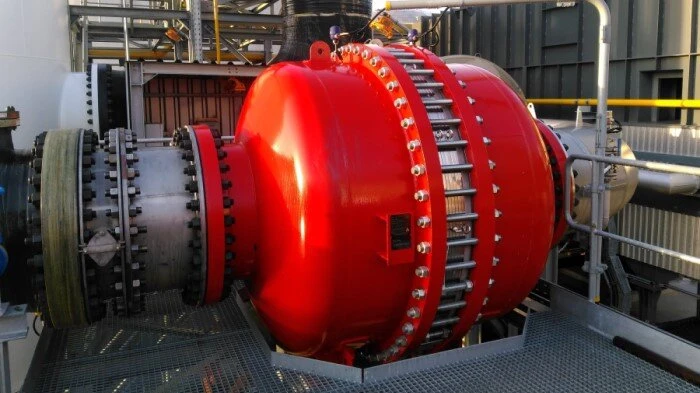 A large red industrial pressure-relief device is bolted into a pipeline on a metal platform at an industrial facility.