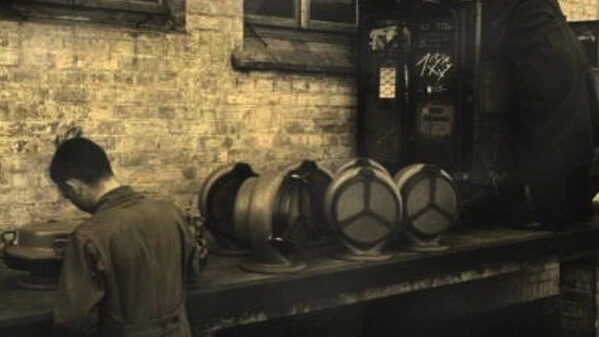 A vintage workshop scene shows workers at a long bench with several large cylindrical metal parts lined up against a brick wall.