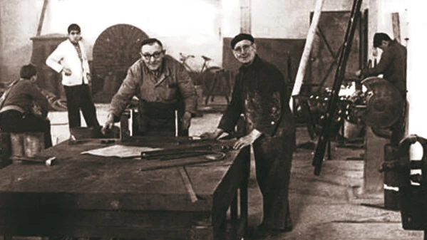 A vintage black-and-white workshop photo shows several workers gathered around a large worktable with tools and metal parts.