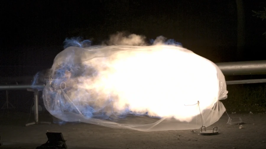 Nighttime industrial explosion test showing a large, bright fireball expanding inside a translucent protective enclosure connected to a pipeline.