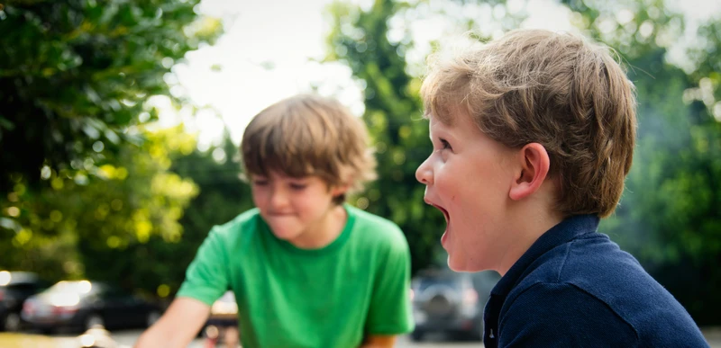Zwei Jungen spielen draußen in einem kleinen rosa aufblasbaren Planschbecken; im Vordergrund lacht einer der Jungen.