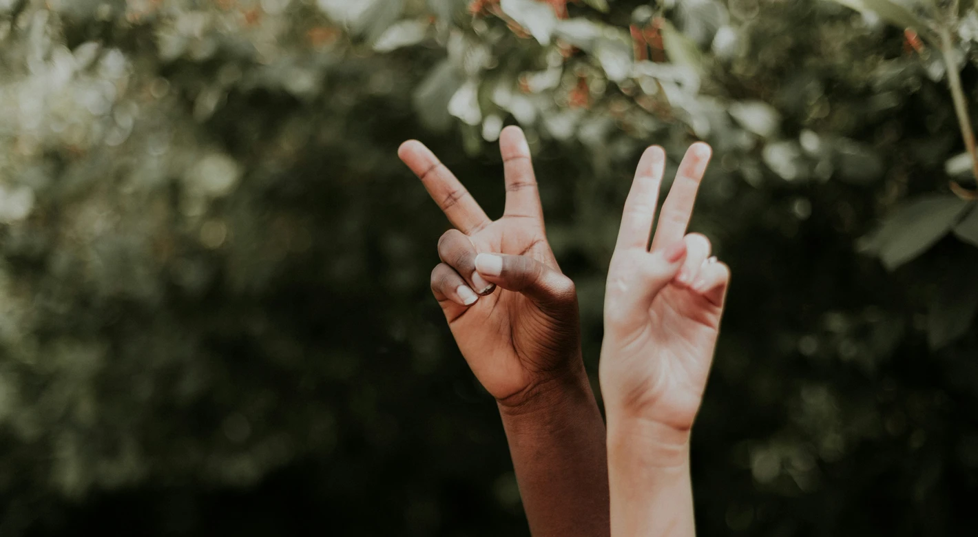 Dos manos levantadas, de distintos tonos de piel, forman el signo de la paz sobre un fondo verde difuminado y frondoso.
