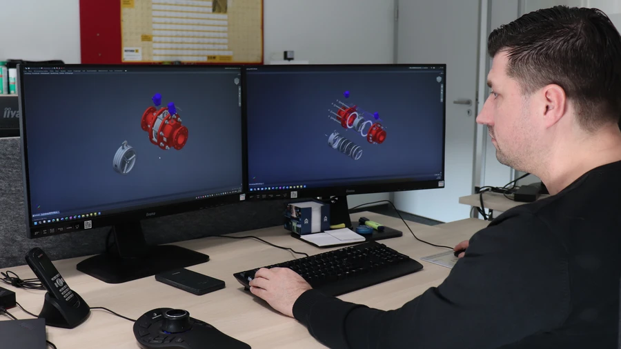 A man works at a desk with dual monitors displaying 3D CAD models of a mechanical assembly, using a keyboard and control device.