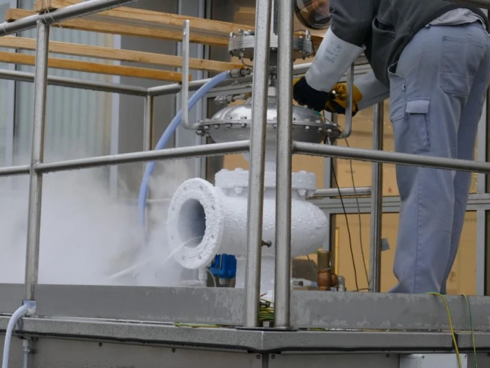 Ein Techniker justiert ein mit Frost bedecktes Industrieventil auf einer erhöhten Plattform, während während der Prüfung kalter Dampf aus dem angeschlossenen Rohr austritt.