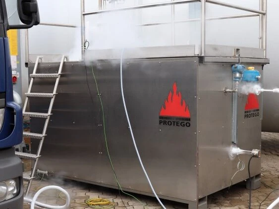 Industrieller Testbehälter aus Edelstahl mit Protego-Logos, Außenverrohrung und einer Leiter, im Freien aufgestellt, mit angeschlossenen Schläuchen und leichtem Dampf.