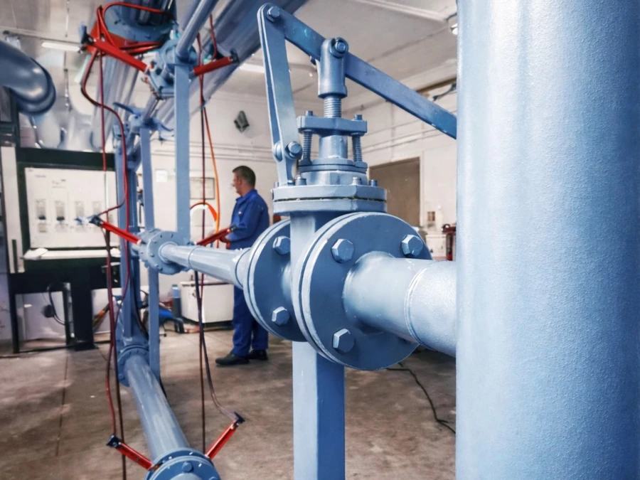 Industrieller Testaufbau in Innenräumen, der eine große blaue Rohrleitung mit einem montierten Ventil und Messgeräten zeigt, die von einem Techniker im Hintergrund überwacht werden.