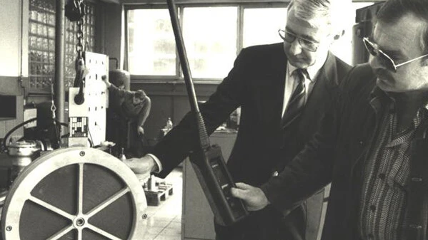 A black-and-white photo shows two men in suits examining industrial equipment beside a large circular device, with one holding a handheld instrument.