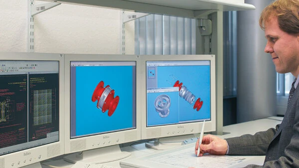 An engineer uses a stylus at a desk while three monitors display exploded-view 3D CAD models of a red mechanical component.