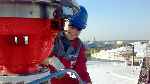 A worker in red coveralls walks through a large industrial plant filled with pipes, valves, and tall processing towers.