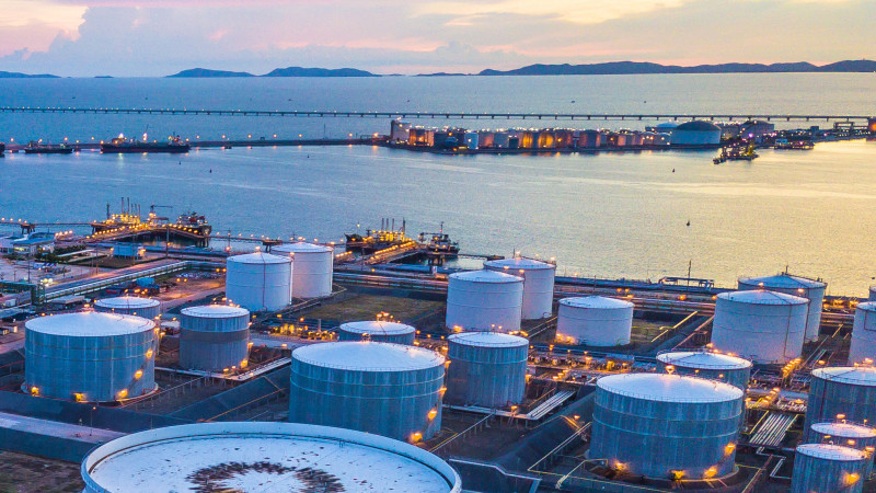 An oil and gas industry complex with large storage tanks near a harbor at dusk